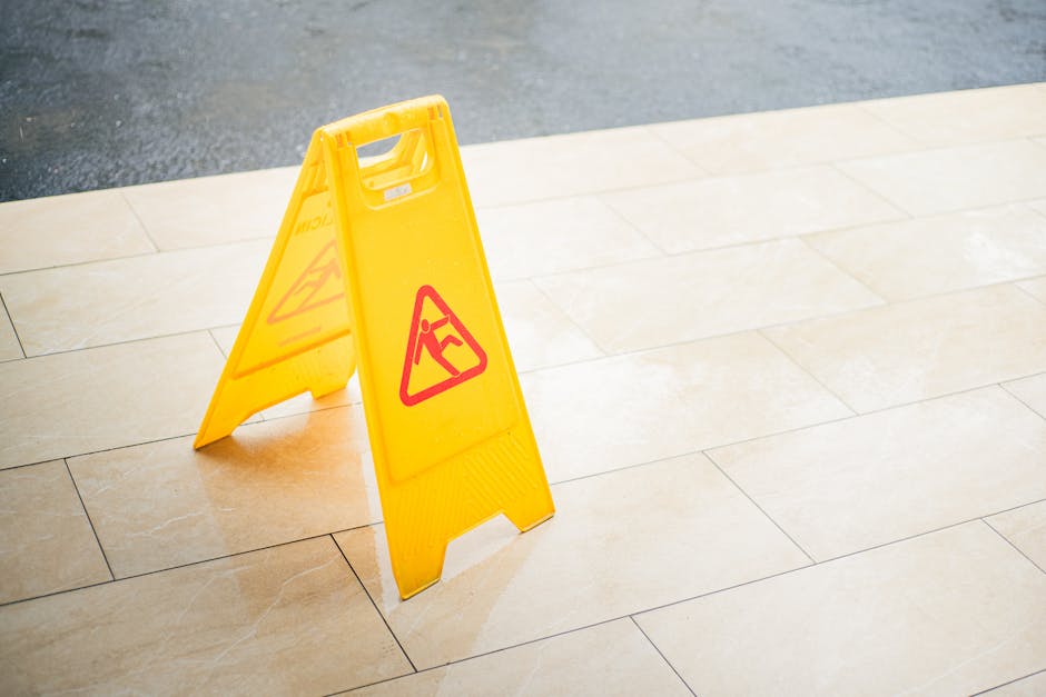 A yellow 'slippery floor' caution sign placed on a light beige tiled indoor floor, indicating a wet or recently cleaned surface. The tiles are smooth, glossy, and clean, reflecting ambient light, with
